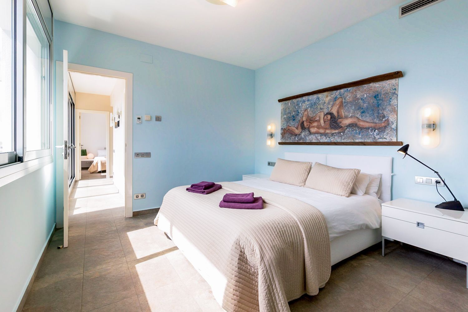 Un dormitorio con una cama, una mesita de noche, un cuadro y una puerta que da a otra habitación.