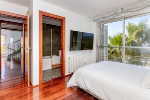 Ein Schlafzimmer mit einem großen Bett, einem Fernseher, einem Fenster mit Blick auf Palmen und einem Badezimmer mit Dusche und Toilette.