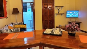 Un salon comprenant une table à manger en bois, un canapé avec des coussins, un téléviseur fixé au mur et une porte donnant sur une salle de bain.