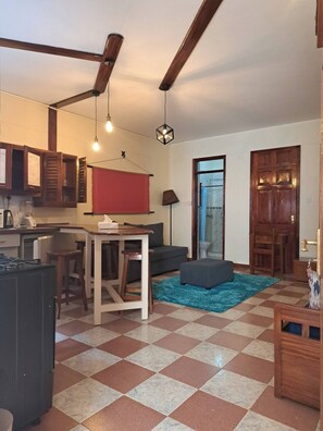 Una sala de estar con piso a cuadros, un sofá, una mesa, y una cocina con armarios, además de una cortina roja.