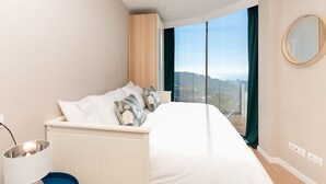 Una camera da letto con un letto, un comodino, una lampada, uno specchio e vista sulla città attraverso ampie finestre.