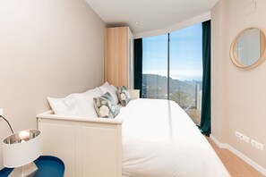 Ein Schlafzimmer mit einem Bett, einem Nachttisch, einer Lampe, einem Spiegel und Blick auf die Stadt durch große Fenster.
