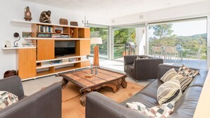 En moderne stue med sofa, sofabord og reol, store vinduer med udsigt til grønne omgivelser.