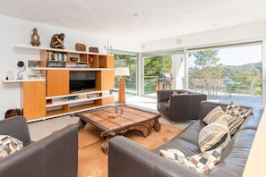 Una sala de estar moderna con sofá, mesa de centro y estantería, ventanas amplias con vistas a zonas verdes.