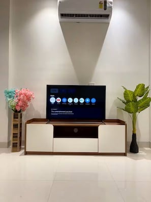 En platt-TV står på ett TV-bord i trä, och bredvid den finns en vas med blommor och en krukväxt.