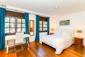 Ein Schlafzimmer mit Bett, Schreibtisch und Stuhl, großen Fenstern mit Vorhängen sowie einem Gemälde an der Wand.
