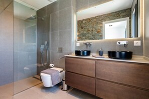 Ein modernes Badezimmer mit Duschkabine aus Glas, einer Wand mit Steinverkleidung und einem Holzbadezimmer-Möbelstück mit zwei schwarzen Waschbecken.