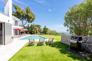 Ein Garten mit Swimmingpool, Liegestühlen, Grill und Blick auf den Ozean.