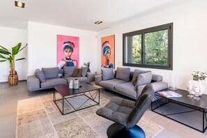 Ett modernt vardagsrum med två soffor, ett soffbord och en fåtölj. På väggen hänger två stora tavlor, och det finns en krukväxt och ett fönster med utsikt över grönområden.