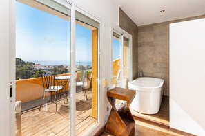 Ein modernes Badezimmer mit einer Badewanne, einem hölzernen Hocker und Meerblick durch eine Glastür.