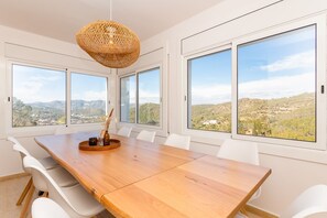 Ein Essbereich mit einem Holztisch, weißen Stühlen und großen Fenstern mit Blick auf eine hügelige Landschaft.