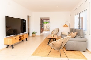 Ein modernes Wohnzimmer mit Flachbildfernseher, grauem Sofa, Korbstuhl, Couchtisch aus Holz und großem Fenster.