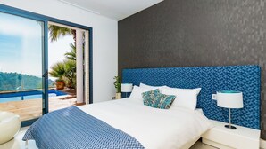 Ein modernes Schlafzimmer mit einem großen Bett, Blick auf einen Pool und Palmen sowie einer dunkel strukturierten Wand.