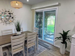 Un coin repas avec une table en bois, des chaises, une plante en pot, un élément décoratif mural et une porte-fenêtre coulissante donnant sur une terrasse.