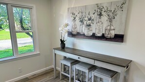 A room with a wooden desk, three stools, a vase with flowers, and a painting of vases on the wall. - Charming 2BR Cottage - Short walk to Missouri Star & Downtown Hamilton (Hamilton)