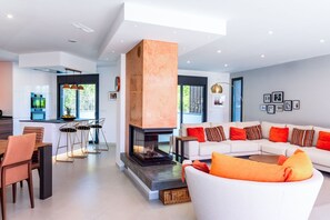 Ein modernes Wohnzimmer mit Kamin, einer Essecke mit Glastisch und einem Sofa mit orangefarbenen Kissen.