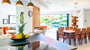 Une cuisine moderne dotée d’un îlot central, d’un coin repas avec des chaises, d’une grande fenêtre offrant une vue sur une piscine et sur la verdure, et d’un éclairage suspendu.