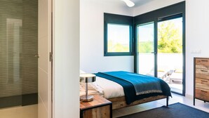 Une chambre à coucher avec un lit, une table de chevet, une lampe, un ventilateur de plafond et une vue sur l’extérieur par une grande fenêtre.