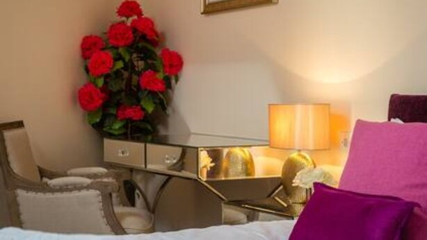 Quarto de hotel com uma cama, uma escrivaninha com abajur, uma cadeira e um vaso grande de flores vermelhas.