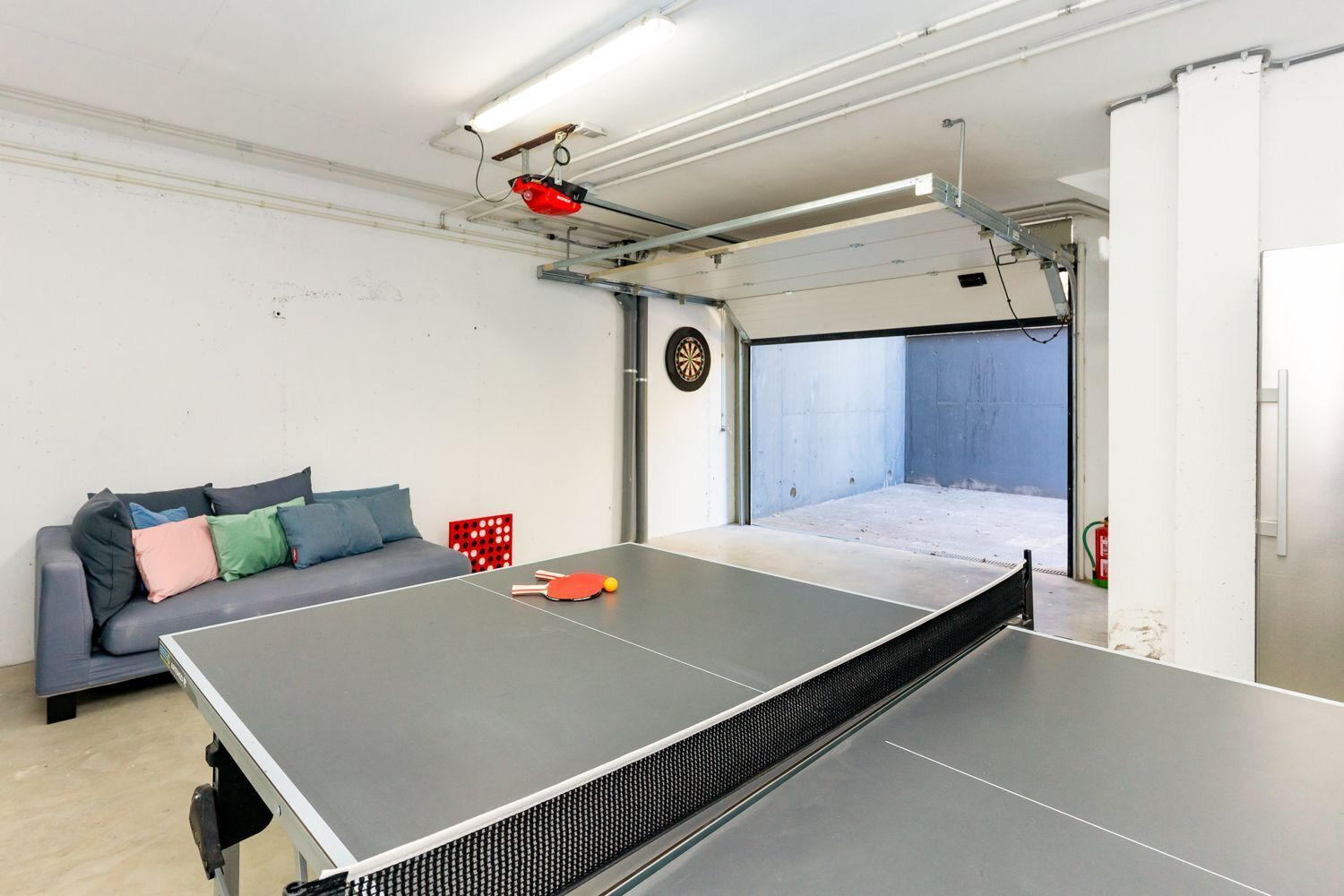 A ping pong table in a room with a sofa, a dartboard, and a door leading to a balcony.