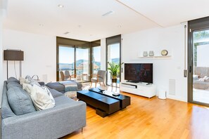 Ein modernes Wohnzimmer mit einer Couch, Sesseln, einem Couchtisch, einem Fernseher und großen Fenstern mit Blick.