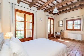 Ein Schlafzimmer mit einem großen Bett, Holzbalkendecke, einem Fenster mit Vorhängen, einem Heizkörper und einem Sessel.