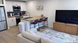 A modern kitchen with white cabinets, a microwave, and a coffee maker. There is a bar area with stools and a large flat-screen TV on a wooden stand.