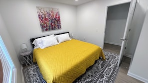 A bedroom with a bed, a nightstand, a lamp, a door, and a patterned rug. - Sewing Serenity B- New 2 BR Home in Hamilton, MO - 3 blocks to Missouri Star (Hamilton)