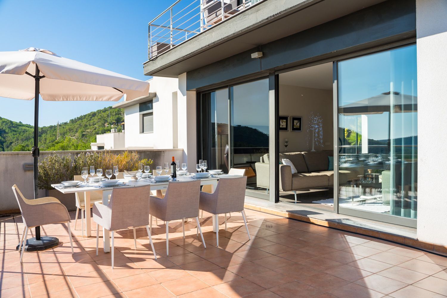 A modern outdoor dining area with a table set for four, a large umbrella, and a view of the interior through glass doors.