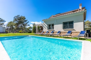 Ein Schwimmbecken mit Liegestühlen, ein weißes Gebäude mit Steinverkleidung und ein klarer, blauer Himmel.