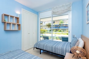 A bedroom with a bed, a bedside table, a lamp, a wardrobe, and a window with a view of a building and greenery. - Apartment Tropicana - Three Bedroom Apartment, Sleeps 6 (Sitges)