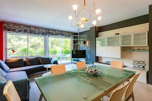 A modern living space with a glass dining table, a sofa, and a television in the background. - Apartment Tropicana - Three Bedroom Apartment, Sleeps 6 (Sitges)