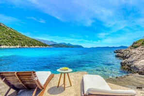 Ein Strand mit klarem, blauem Wasser, einem Holzliegestuhl, einem weißen Liegestuhl und einem kleinen runden Tisch mit einem gelben Gegenstand darauf.