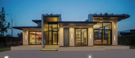 Una casa moderna con grandes ventanales, una entrada iluminada y un dormitorio visible.
