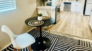 A compact kitchen with white cabinets, a microwave, a stove, and a dishwasher. There is a small dining area with a round table and a white chair. - Stylish Downtown Cottage near Alamodome, Hemisphere & Riverwalk (San Antonio)