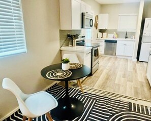 A compact kitchen with white cabinets, a microwave, a stove, and a dishwasher. There is a small dining area with a round table and a white chair. - Stylish Downtown Cottage near Alamodome, Hemisphere & Riverwalk (San Antonio)