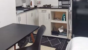 Een kleine keuken met witte kastjes, een zwart aanrecht, een eettafeltje en een zwarte koelkast.