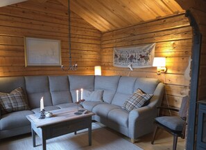 A cozy cabin interior with a wooden ceiling, a grey sofa, a wooden coffee table with candles, and a wall-mounted tapestry. - Mountain Holiday Homes (Undersaker)