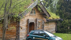Une cabane en rondins avec une voiture garée devant, entourée d’arbres.