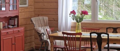 A wooden cabin interior with a dining table, chairs, a rocking chair, a pink cabinet, and a window with a view of trees.