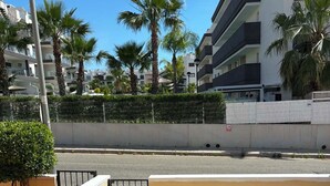 Un balcon avec une balustrade en métal, une allée pavée et une vue sur une rue bordée de palmiers et d’immeubles.