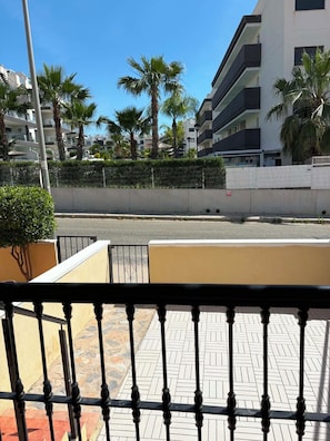 Ein Balkon mit Metallgeländer, ein gepflasterter Weg und Blick auf eine Straße mit Palmen und Gebäuden.