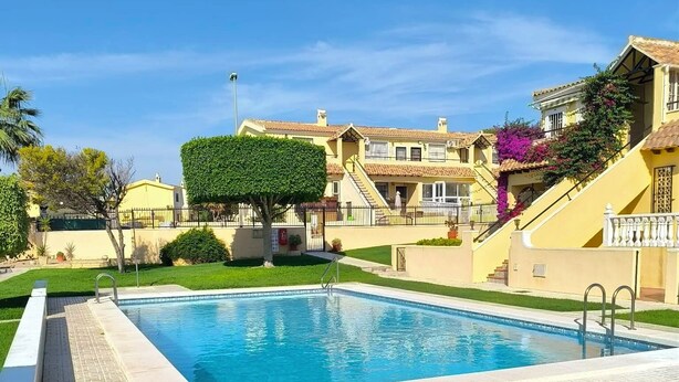 Une piscine dotée d’une terrasse carrelée, entourée de pelouse bien entretenue et bordée d’une rangée d’immeubles avec des balcons.