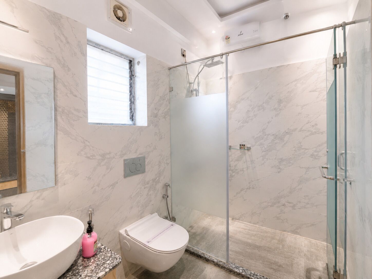 Un baño moderno con suelo de mármol, ducha con cerramiento de vidrio, inodoro blanco y lavamanos con dispensador de jabón rosa.