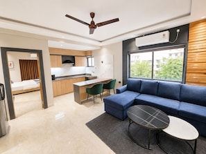 A modern living room with a blue sofa, a small round table, and a kitchen area with a wooden countertop and a built-in oven. - The Bay Suites - NIS (Barasat)