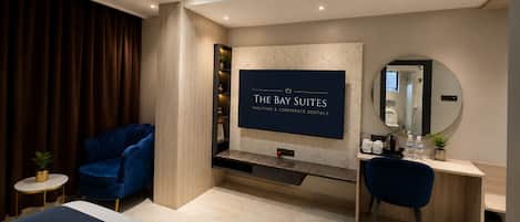 A modern hotel room with a large TV screen displaying 'THE BAY SUITES' branding, a desk with a mirror, a blue chair, and a bed with a dark blue headboard.