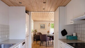 Ett kök med vita skåp, ett träpanelat tak, en matplats med stolar och ett bord samt en förvaringslåda märkt med texten ”FÖRVARING”.