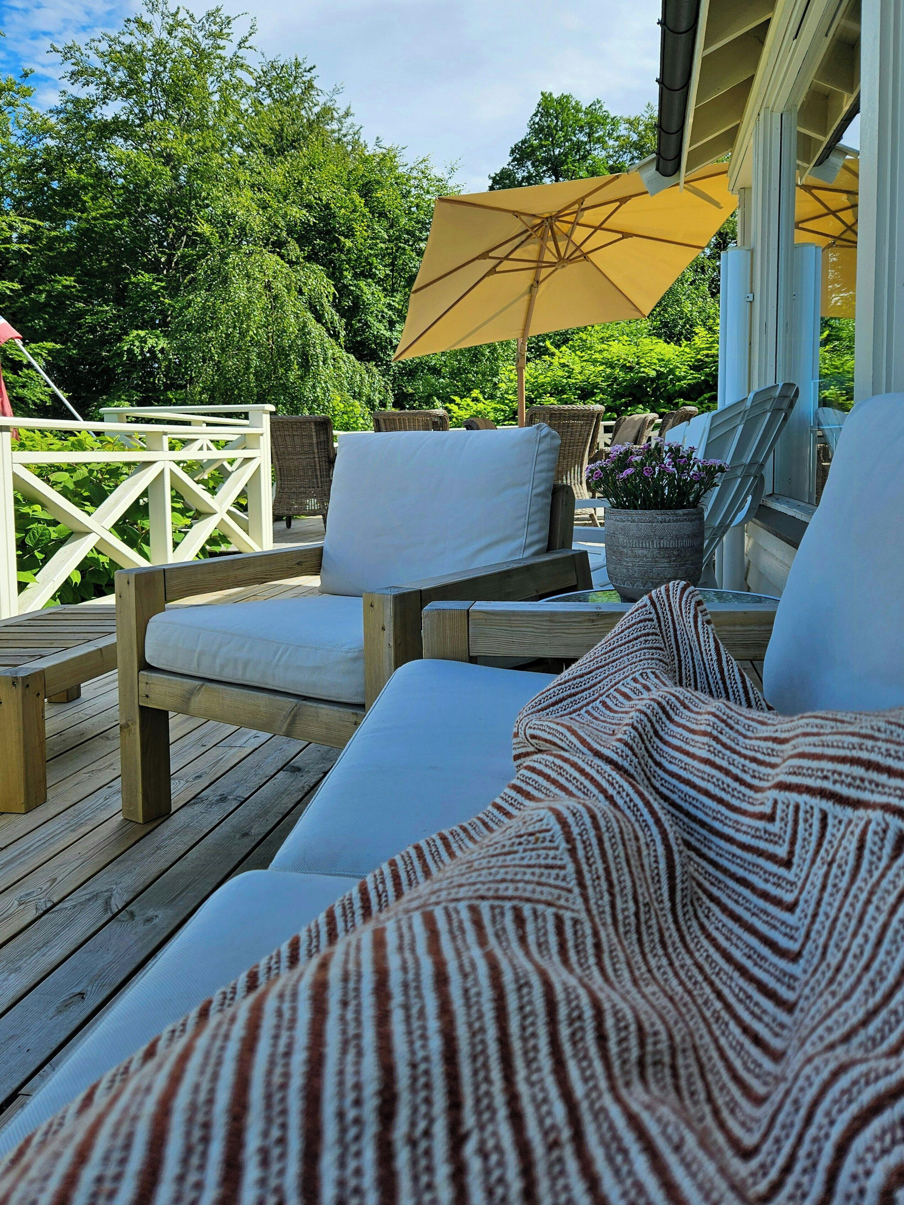 Een houten terras met buitenmeubilair, waaronder een bank en stoelen, en een grote parasol die schaduw biedt.