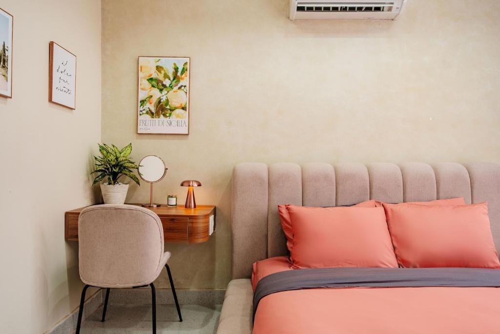Ein Schlafzimmer mit einem Bett, einem Schreibtisch mit Stuhl, einer Topfpflanze, einer Wand-Klimaanlage und gerahmten Kunstwerken.