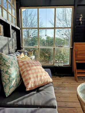 A cozy room with a wooden floor, a sofa with patterned cushions, a large window with a view of trees, and hanging lanterns. - Personal house with direct access to the beach (Sjællands Odde)